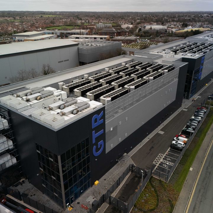 Oblique aerial view of the three GB One buildings with equipment on their roofs. A road and parking spaces run along the right.